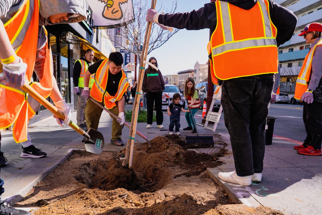 The image shows a group of people planting a tree on a city sidewalk. Several individuals, wearing bright orange safety vests, are actively participating in the planting process. Two people are holding shovels and appear to be digging a hole, while another person steadies the tree. In the background, a woman and two children observe the activity. The scene is set on a bustling city street with storefronts and a few bystanders in the background. The day appears sunny, casting clear shadows on the sidewalk.