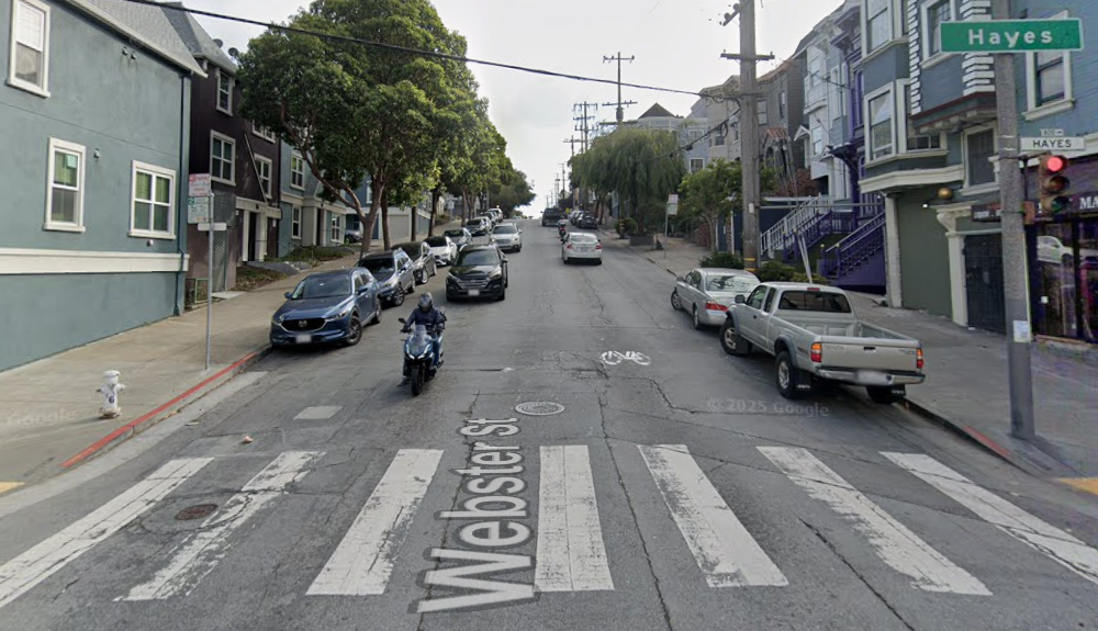 Street level view of the Webster Street and Hayes Street intersection looking south toward Fell Street. Depicted are the roadway with vehicles street parked on either side, as well as the buildings that line this block of Webster Street.