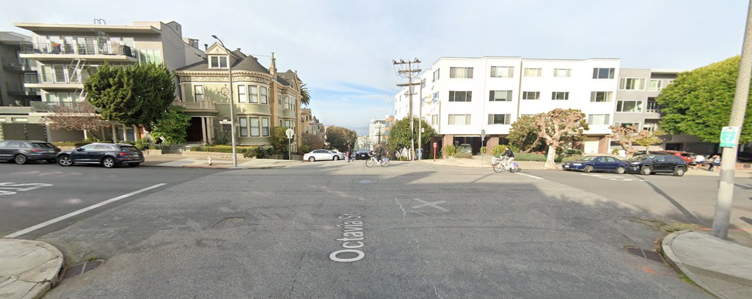 Street level view of the Octavia Street and Pacific Avenue intersection. A wide-angle view of the roadway, sidewalks and adjacent buildings is shown, facing north. A cyclist rides through the intersection heading west along Pacific Avenue.