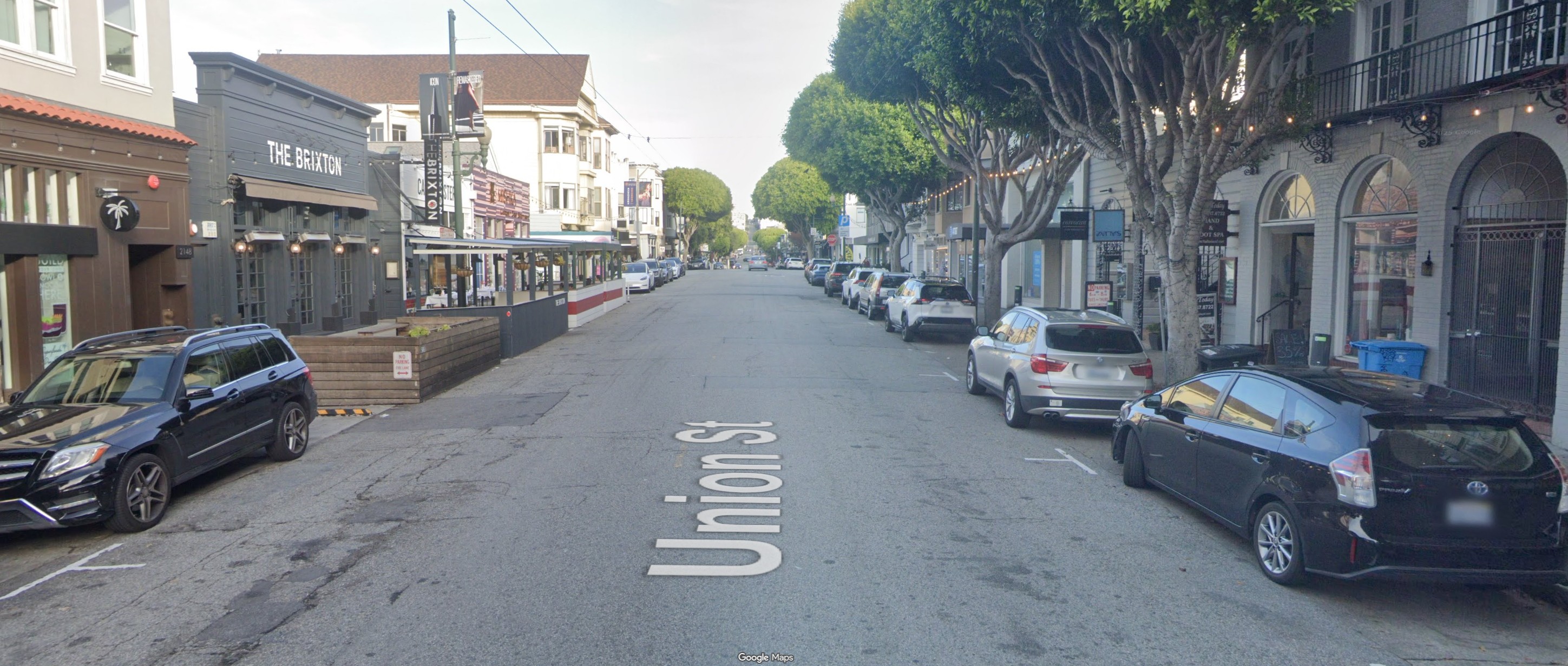 Street level view of Union Street. Visible is the roadway, which is cracked and uneven in places, especially in the left hand lane. Vehicles are parked on either side of the street, and there are outdoor dining parklets in front of some of the neighboring businesses.