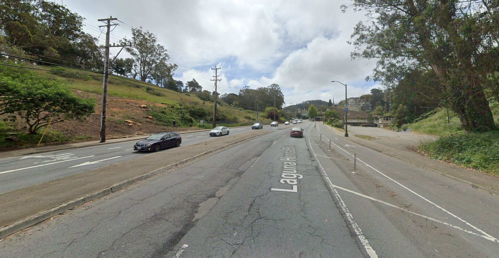 Street level view of Laguna Honda Boulevard depicting multiple vehicles, roadway lanes and a two-way bicycle lane. On either side of the roadway are trees, shrubs and a sloping hillside on the left-hand side.