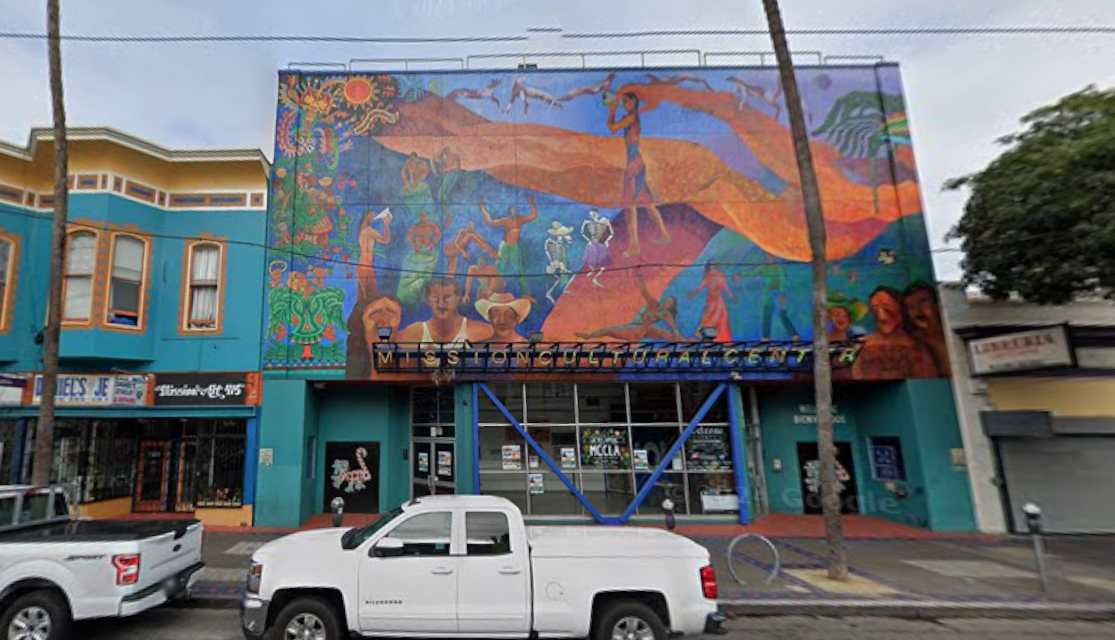 Street level view of the Mission Cultural Center, showing the building's entryway, exterior windows on the ground floor and a large, multi-story mural painted on the upper facade.